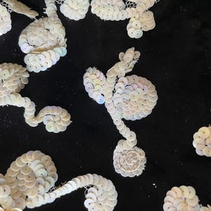 Op de afbeelding: Een zwart stoffen oppervlak versierd met meerdere witte iriserende lovertjes in bloemmotieven. Elk motief bestaat uit kleine, ronde, lichtreflecterende lovertjes die in organische, wervelende patronen zijn gerangschikt voor een textuur.