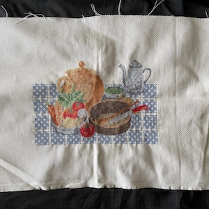 Peut inclure: Broderie au point de croix représentant une nature morte de cuisine. Une théière marron, une théière en argent et un bol de nourriture sont disposés sur une nappe à carreaux bleus et blancs. Des tomates rouges et des herbes vertes sont également visibles.