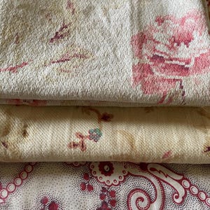 May include: Three folded pieces of fabric stacked vertically. The top layer is cream with a large pink floral pattern. The middle layer is light beige with small floral sprigs. The bottom layer is off-white with a detailed red paisley print.