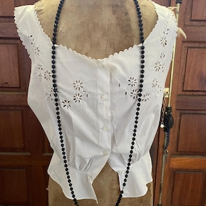 Può includere: Una camicetta bianca traforata con ritagli floreali e un'abbottonatura frontale è esposta su un manichino vintage. Una lunga collana di perline nere pende sopra la camicetta. La camicetta ha uno scollo smerlato e maniche corte.