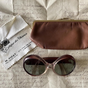 May include: Vintage sunglasses with a brown frame and green lenses, beside a brown leather case and a cleaning cloth with the text "Optique des Carmes". The items are on paper with handwritten text.