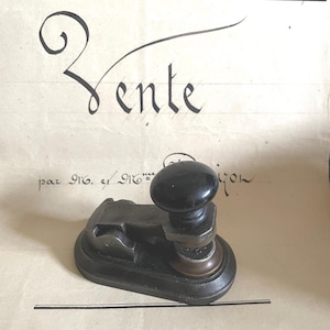 Op de afbeelding: Een vintage handmatige reliëfstempel van zwart metaal met een afgeronde handgreep en een stevige voet. Het apparaat staat op een crèmekleurig document met handgeschreven Franse tekst, inclusief de datum 2 Avril 1876.