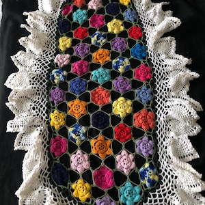 Könnte beinhalten: Handgefertigter, ovaler Tischläufer aus Häkelarbeit mit farbenfrohem Blumendesign. Die Mitte ist mit leuchtenden, mehrfarbigen Blumen in Rot-, Orange-, Gelb-, Rosa-, Lila- und Blautönen gefüllt. Der Rand ist eine zarte weiße Spitze.