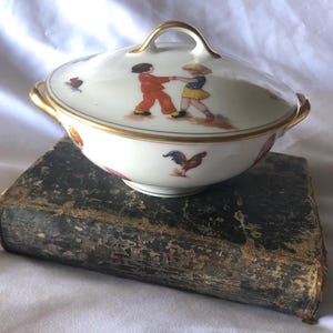 Könnte beinhalten: Eine weiße Porzellan-Zuckerdose mit Deckel, verziert mit einem handgemalten Design von zwei tanzenden Kindern mit einem Hahn. Die Dose hat einen goldenen Rand und steht auf einem dunkelbraunen Buch.