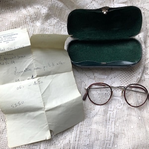 May include: A handwritten note with the text "Dr. Michel Coutris" and "Lunette avec pr t k aller" along with a price of £2.50. A green glasses case with a silver clasp and a pair of brown-rimmed glasses are also pictured.