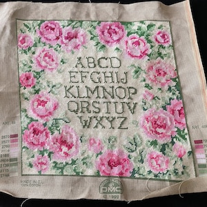 May include: Needlepoint canvas featuring a floral design with pink roses and green leaves. The letters A through Z are stitched in a dark green font. The canvas is on a beige background with a DMC logo.