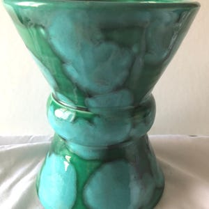 Könnte beinhalten: Eine türkisfarbene und grüne Keramikvase mit einer ungewöhnlichen Sanduhrform. Die Vase hat eine breite Ober- und Unterseite, die durch einen schmaleren, abgerundeten Mittelteil verbunden sind. Die Glasur hat ein gesprenkeltes, marmoriertes Aussehen.