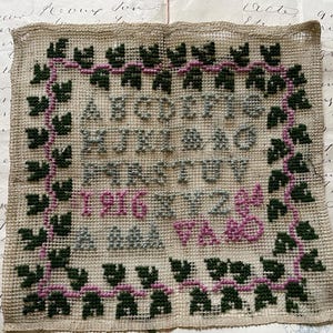 以下が含まれることがあります： 1916年のアルファベットが刺繍されたヴィンテージのニードルポイントサンプラー。ベージュの背景に緑、ピンク、グレーのクロスステッチの文字と、葉とブドウの木のボーダーがあります。