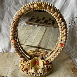 May include: Oval vanity mirror decorated with seashells and faux pearls. The frame is adorned with various seashells, with a base also embellished with shells. The mirror reflects a vintage aesthetic, perfect for coastal or nautical decor.