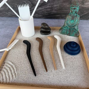 May include: A miniature zen garden set with a wooden frame, sand, and various accessories. Includes a small Buddha statue, black stones, rakes, and decorative hair sticks in black, brown, and white. A dark blue, layered disc is also present.