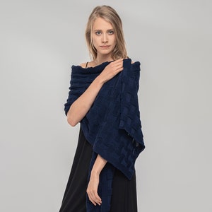 May include: A woman wearing a black dress and black leggings with a navy blue knitted shawl draped around her shoulders. She is wearing black ankle boots.