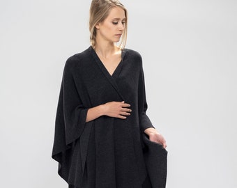 May include: A charcoal grey knit poncho with a draped design. The poncho is worn over black leggings and black ankle boots.