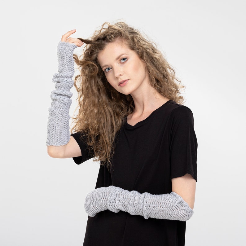 May include: A woman with curly blonde hair is wearing a black t-shirt and gray fingerless gloves. The gloves are knitted and have a textured pattern.
