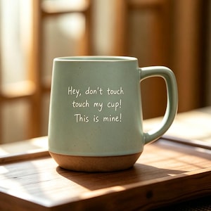 May include: A light green ceramic mug with a speckled finish and a brown unglazed base. The front of the mug features the white printed text: Hey, don't touch touch my cup! This is mine! The mug has a single handle on the right side.