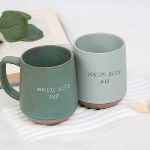 Può includere: Due tazze in ceramica, una verde acqua e una azzurra, con le scritte "WORLD'S BEST DAD" e "WORLD'S BEST MUM". Le tazze sono su un panno bianco a righe, con un libro e del verde sullo sfondo.