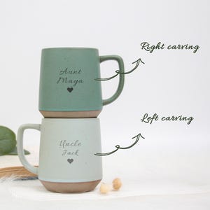 May include: Two ceramic mugs, one stacked on the other. The top mug is a muted green with the text "Aunt Maya" and a heart. The bottom mug is light blue with "Uncle Jack" and a heart. Both mugs have a brown base and a curved handle.