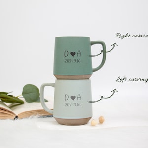 May include: Two ceramic mugs, one sage green and one light blue, stacked on top of each other. Each mug has a handle and a base with a brown color. The mugs are engraved with a heart symbol, the letter "D", the letter "A", and the date "2024.9.16".