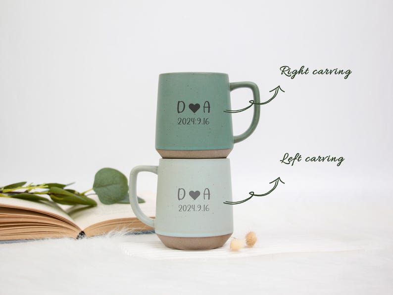 May include: Two ceramic mugs, one sage green and one light blue, stacked on top of each other. Each mug has a handle and a base with a brown color. The mugs are engraved with a heart symbol, the letter "D", the letter "A", and the date "2024.9.16".