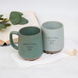 May include: Two ceramic mugs, one teal and one light blue, with a textured finish and a brown base. Each mug has the inscription "Hudson ♡ Aria 5 years 2025". The mugs are on a white surface with a book and greenery in the background.