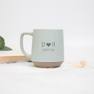 May include: A light blue ceramic mug with a brown base and a curved handle. The mug features the initials "D A" with a heart symbol and the date "2024.9.16" printed in gray. The mug is set against a white background.
