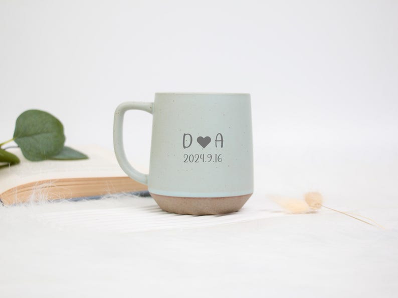 May include: A light blue ceramic mug with a brown base and a curved handle. The mug features the initials "D A" with a heart symbol and the date "2024.9.16" printed in gray. The mug is set against a white background.
