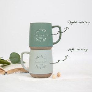 May include: Two ceramic mugs, one sage green and one light blue, stacked. Each mug has a brown base and a curved handle. The mugs are decorated with a floral design and the text "David Emily 2026".