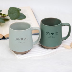 Puede incluir: Dos tazas de cerámica, una azul claro y otra verde, con base marrón. Cada taza tiene un asa y está inscrita con iniciales, un corazón y la fecha 2016.9.29. Las tazas están sobre una superficie blanca.