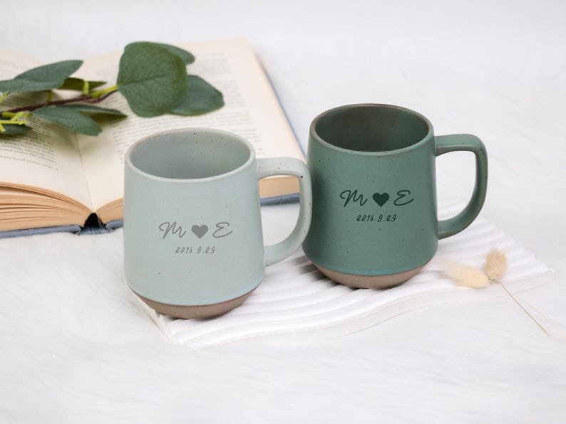 May include: Two ceramic mugs, one light blue and one green, with a brown base. Each mug has a handle and is inscribed with initials, a heart, and the date 2016.9.29. The mugs are on a white surface.