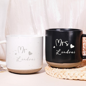 May include: A pair of ceramic mugs, one white and one black, with a speckled design. The white mug has "Mr. Lendrai" printed on it, and the black mug has "Mrs. Lendrai" printed on it. Both mugs have a brown base and a heart symbol.
