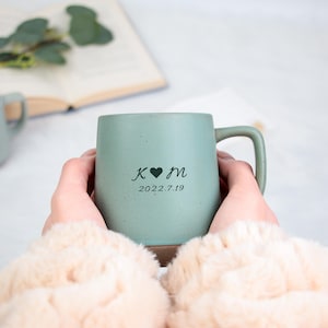 May include: A light green ceramic mug with a handle, personalized with initials, a heart, and the date 2022.7.19. The mug is held by a person wearing a cream-colored sweater. Another mug is visible in the background.