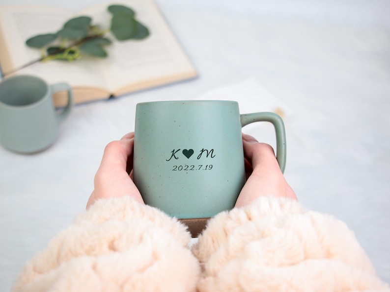 May include: A light green ceramic mug with a handle, personalized with initials, a heart, and the date 2022.7.19. The mug is held by a person wearing a cream-colored sweater. Another mug is visible in the background.