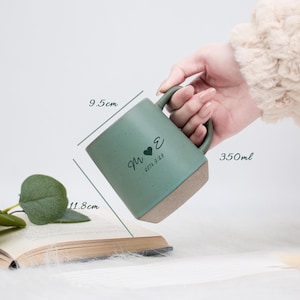 May include: A teal ceramic mug with a brown base, held by a hand. The mug has a heart and initials design, with the date 2016.9.29. It measures 9.5cm x 11.8cm and holds 350ml. The mug is placed near an open book.