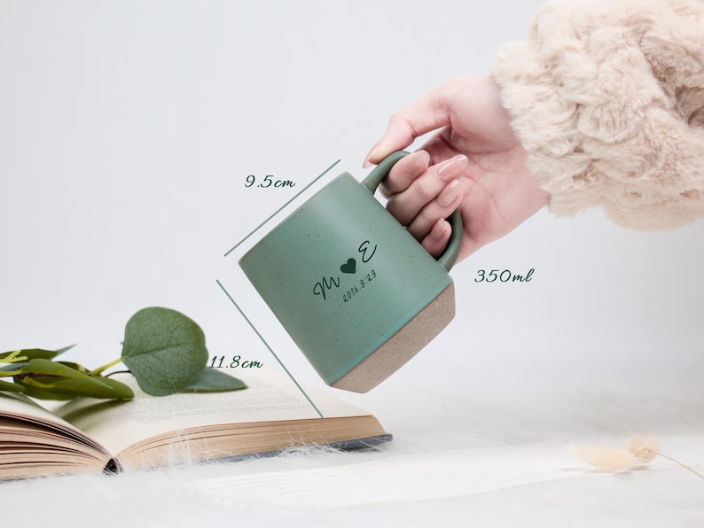May include: A teal ceramic mug with a brown base, held by a hand. The mug has a heart and initials design, with the date 2016.9.29. It measures 9.5cm x 11.8cm and holds 350ml. The mug is placed near an open book.