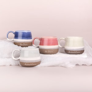 May include: Four ceramic mugs in blue, pink, cream, and light green. Each mug has a white band and a brown base. The mugs have white handles and feature personalized text. The mugs are on a white surface against a soft pink background.