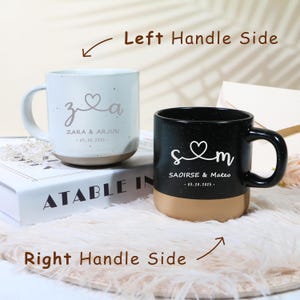 Puede incluir: Dos tazas de cerámica con texto personalizado. Una es blanca con acabado moteado, la otra es negra. Ambas tienen base beige y asa negra. La taza blanca dice "Zara & Arjun" y la negra "Saoirse & Mateo" con una fecha.