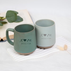 May include: Two ceramic mugs, one with a handle and one without, in shades of green and gray. Each mug features a personalized design with initials, a heart symbol, and a date. The mugs rest on a white surface.