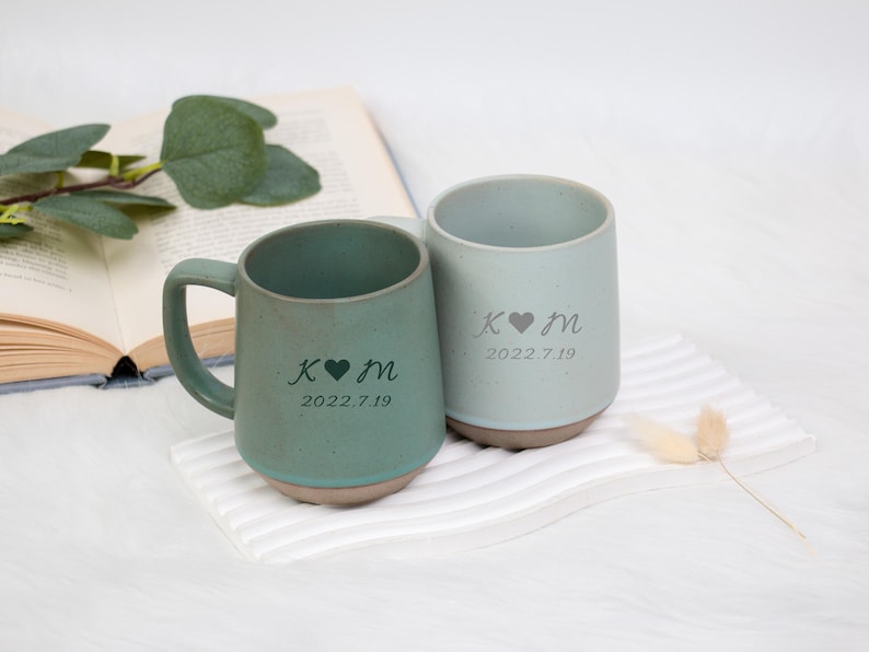 May include: Two ceramic mugs, one with a handle and one without, in shades of green and gray. Each mug features a personalized design with initials, a heart symbol, and a date. The mugs rest on a white surface.