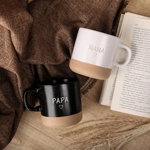 Pode incluir: Duas canecas de cerâmica com um esmalte salpicado. A caneca à esquerda é preta com um fundo bege e diz "PAPA" em letras brancas com um coração. A caneca à direita é branca com um fundo bege e diz "MAMA" em letras brancas com um coração.