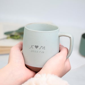 May include: A light blue ceramic mug with a brown base, held in hands. The mug has a handle and is engraved with initials, a heart symbol, and the date 2022.7.19. Another green mug is visible in the background.