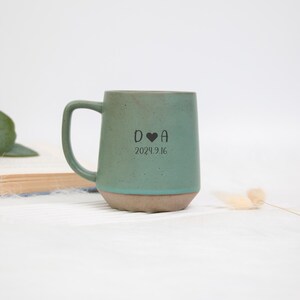 May include: A teal ceramic mug with a rounded handle and a light brown base. The mug features the initials "D ❤️ A" and the date "2024.9.16" printed in black. The mug is set against a white background.