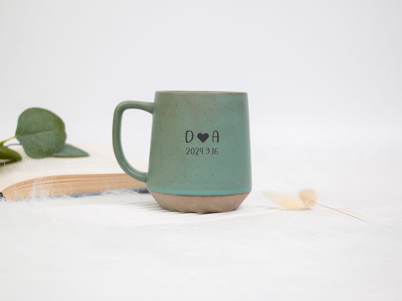 May include: A teal ceramic mug with a rounded handle and a light brown base. The mug features the initials "D ❤️ A" and the date "2024.9.16" printed in black. The mug is set against a white background.