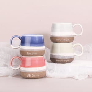 May include: Four ceramic mugs in blue, pink, and green, stacked in pairs. Each mug has a white stripe and a brown base. The mugs are personalised with names such as "Mr. Davies" and "Henry & Abigail."