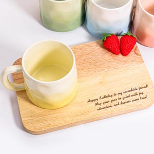 May include: A ceramic mug with a yellow and white gradient sits on a wooden cutting board. The board has a message engraved on it, and two red strawberries are placed nearby. Other mugs in shades of green, blue, and pink are in the background.