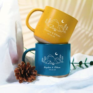 Puede incluir: Dos tazas de cerámica, una amarilla mostaza y otra verde azulado, apiladas una encima de la otra. Cada taza presenta un diseño blanco de montaña y árboles con una tienda de campaña y una luna. Las tazas están personalizadas con los nombres "Sophia & Ethan" y la fecha "05.22.25".