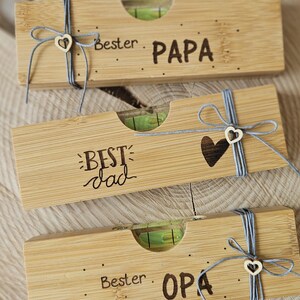 Könnte beinhalten: Drei Holz-Wasserwaagen mit graviertem Text und dekorativem Garn. Die Wasserwaagen lesen "Bester PAPA", "BEST dad" und "Bester OPA". Jede Wasserwaage hat ein herzförmiges Detail und eine Libelle.