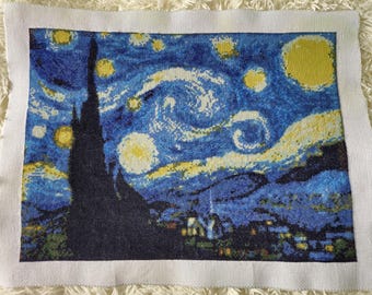 May include: A cross-stitch embroidery of Vincent van Gogh's famous painting, "Starry Night". The artwork depicts a swirling night sky with a bright yellow moon and stars, a dark cypress tree, and a small village in the distance.