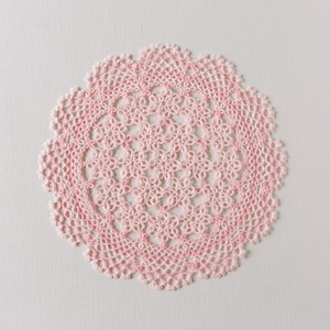 May include: A delicate pink crocheted doily with a circular design. The doily is made of fine thread and has a delicate, lacy appearance.