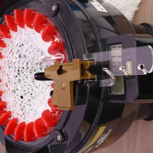 Op de afbeelding: Een close-up van een witte breimachine met rode naalden en een bruine plastic garengeleider. De breimachine heeft een digitale teller met een "CLEAR"-knop.