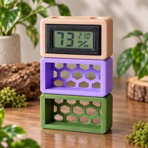 Puede incluir: Una pila de tres bloques rectangulares. El bloque superior es de color melocotón y contiene una pantalla digital que muestra 73% y 21,0°C. El bloque central es morado y el inferior es verde oliva, ambos con un patrón de panal.