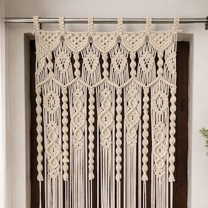 以下が含まれることがあります： クリーム色のマクラメドアカーテン。カーテンは、上部と垂直ストランドに沿って複雑な織り模様が特徴です。デザインには幾何学的な形状と結び目のディテールが含まれており、ボヘミアンスタイルの美学を作り出しています。カーテンは濃い茶色のドアの前に吊るされています。
